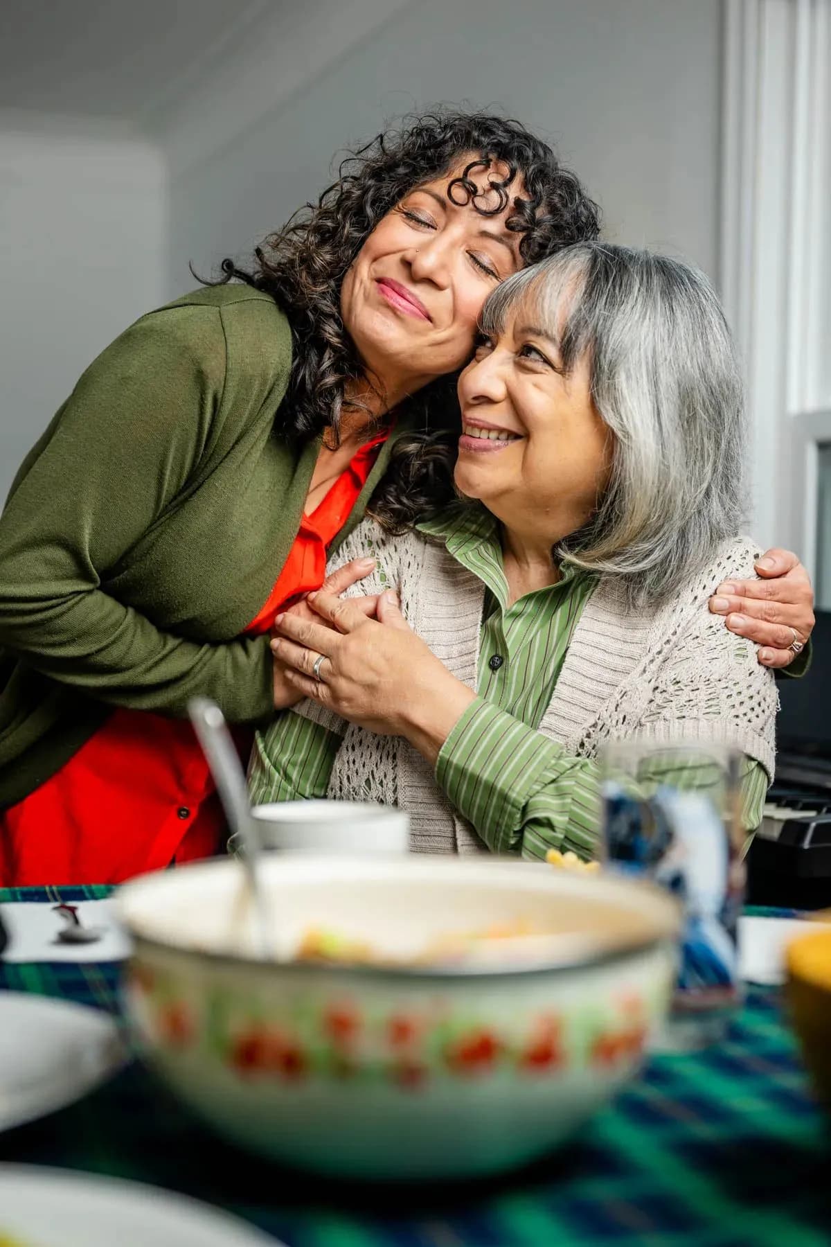 A woman with curly hair hugs a smiling older woman with grey hair