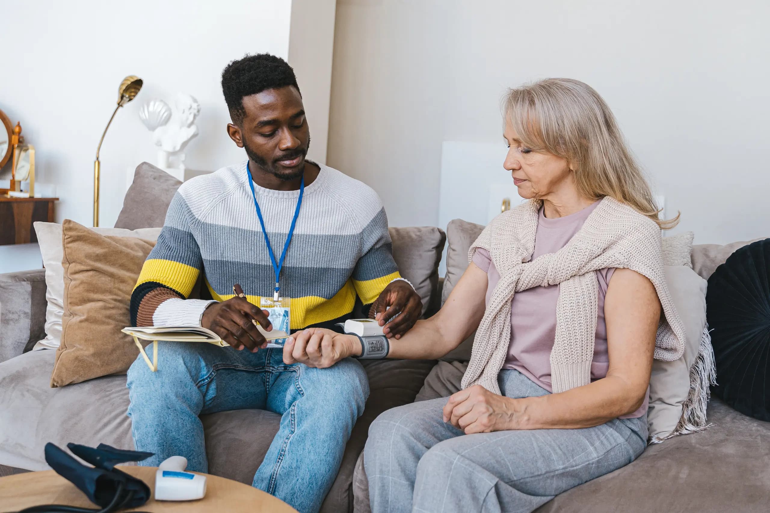 A career checks an older woman's blood pressure as they sit together on a sofa