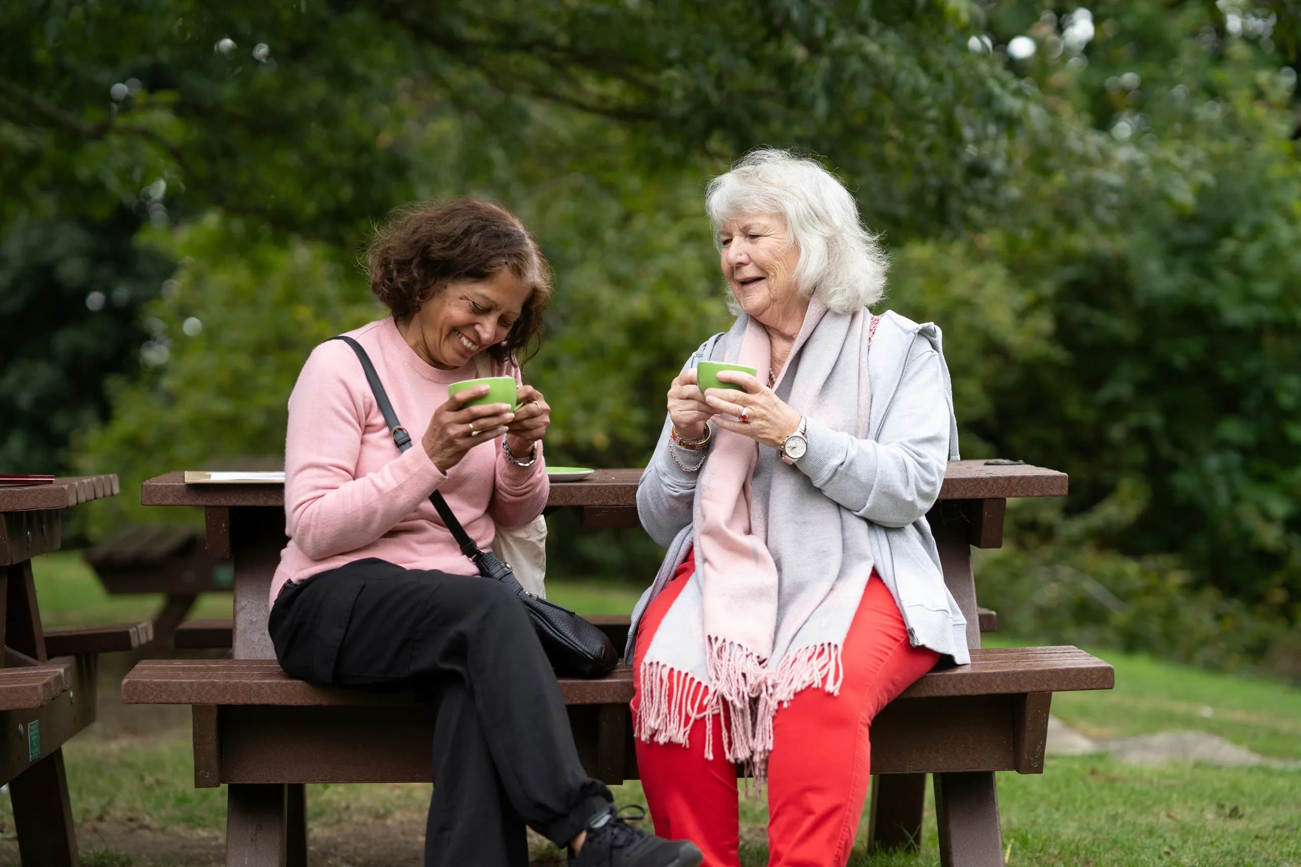 Two older women laughing together over cups of tea at a picnic bench in a garden