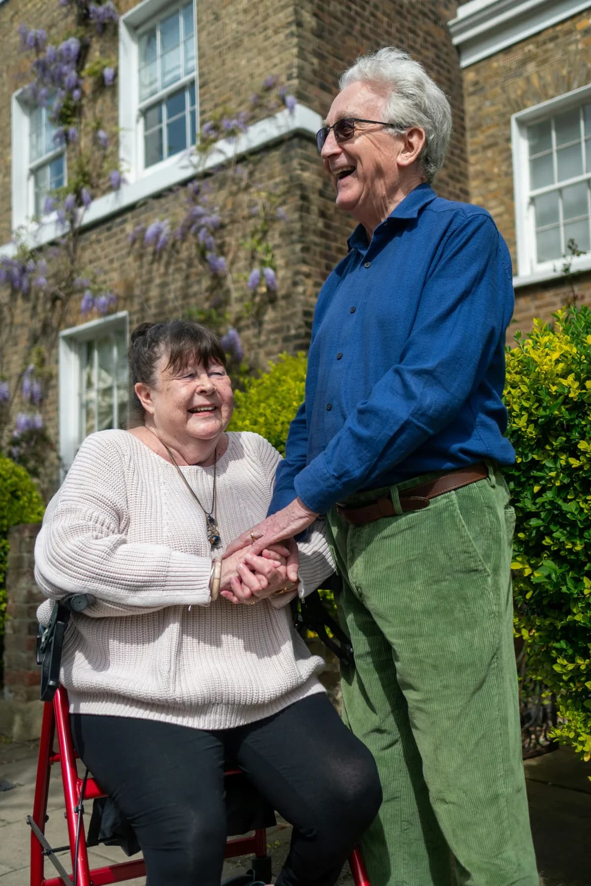 An older woman seated on her walker, laughing and holding hands with a smiling older man outside a brick house